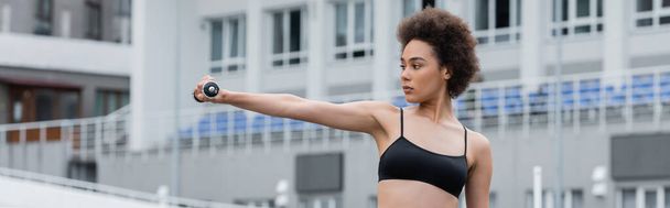 young african american woman in black sports bra training with dumbbell in outstretched hand, banner - Foto, immagini