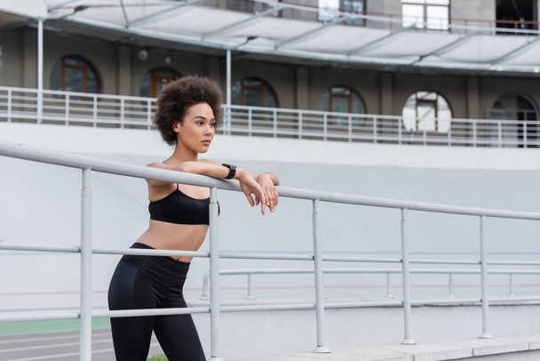 curly african american woman in black leggings and sports bra looking away near fence - Foto, Imagen
