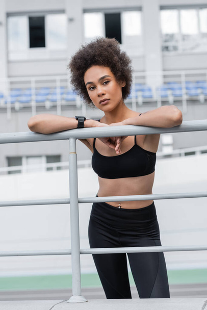 slender african american woman in sportswear and fitness tracker looking at camera near fence - Foto, immagini