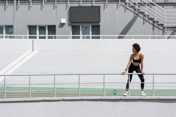 full length of african american woman in black sportswear standing near sports bottle on city stadium - Fotoğraf, Görsel