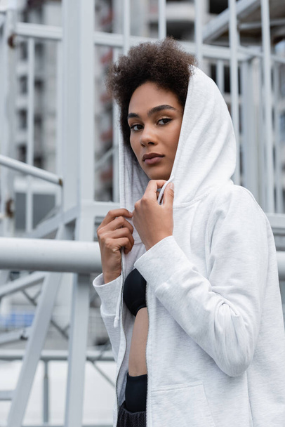 brunette african american woman in white hoodie looking at camera on urban stadium - Foto, Imagen
