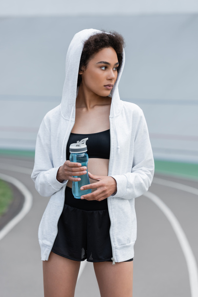 νεαρή Αφροαμερικανή με μαύρα αθλητικά και άσπρη κουκούλα που κρατάει αθλητικό μπουκάλι και κοιτάζει αλλού. - Φωτογραφία, εικόνα