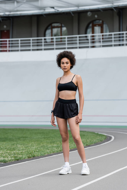 full length of african american woman in black sports bra and shorts looking at camera on stadium - Zdjęcie, obraz