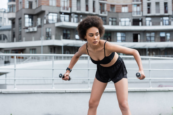 African american sportswoman exercising with dumbbells on blurred stadium  - 写真・画像