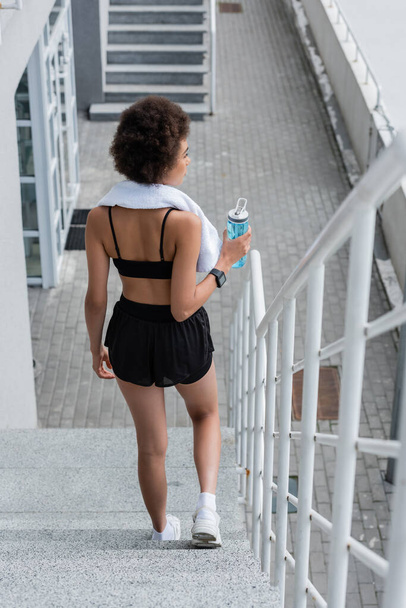 Вид сбоку на африканскую женщину с игрушкой и спортивной бутылкой, идущую по лестнице на стадионе  - Фото, изображение