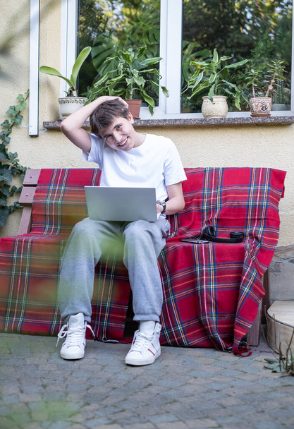 Усміхнений підліток з ноутбуком на лавці біля будинку. Технологічне вивчення, дистанційний онлайн-клас з відеозв'язку, віртуальний урок, спілкування в соціальних мережах, вивчення шкільних матеріалів
 - Фото, зображення