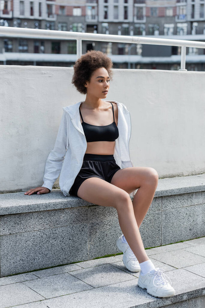 Young african american sportswoman in sports jacket and top sitting on stadium  - Foto, Imagem