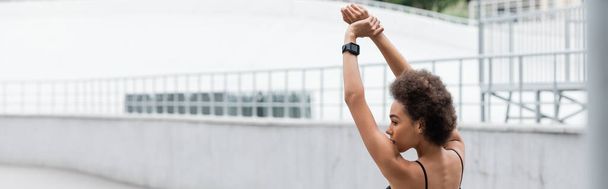 Πλευρική άποψη του Αφροαμερικανού αθλητή με έξυπνο ρολόι που εκτείνεται σε γήπεδο, πανό  - Φωτογραφία, εικόνα
