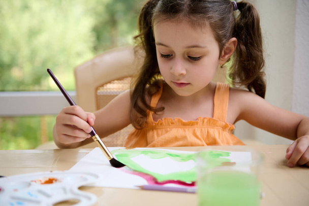 European cute little girl smiles, holding paintbrush and drawing picture while learning watercolour painting class. Drawing. Peinture. Creativity. Hobby and leisure. Kids entertainment and development - Fotoğraf, Görsel
