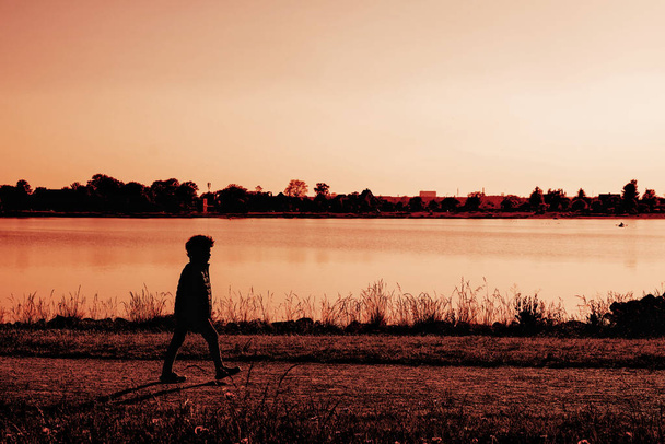 Silhouette of boy near the lake, a lonely child walks along the river at sunset, summer landscape. - Fotografie, Obrázek