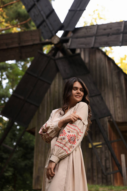 Beautiful woman wearing embroidered dress near old wooden mill in countryside. Ukrainian national clothes - Zdjęcie, obraz