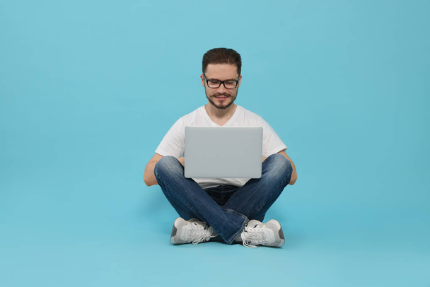 Smiling man sitting and using laptop on light blue background - Фото, изображение
