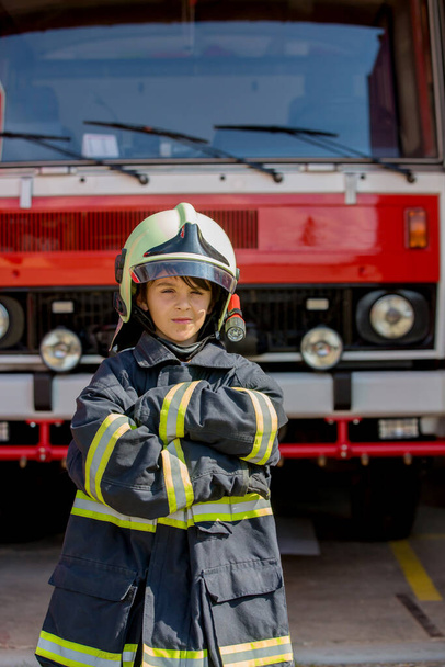 Child, cute boy, dressed in fire fighers cloths in a fire station with fire truck, childs dream - Valokuva, kuva