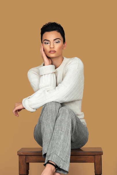 Portrait of a beautiful young White woman with short black hair, eye makeup and lipstick sitting by herself on a table top inside a studio with a pecan background wearing a beige pullover and grey pants. - Foto, Imagem