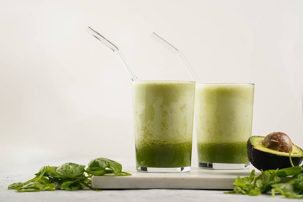 A glass of green healthy detox smoothie with spinach, matcha powder and a glass straw, fresh spinach and avocado on marble board on grey background - Zdjęcie, obraz