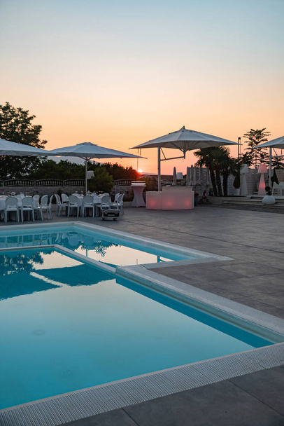 Blue swimming pool in a spa hotel in southern Italy at sunset. Set round white tables for the party - Zdjęcie, obraz