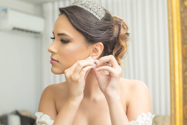 Молодая женщина в свадебном платье, перед зеркалом, надевает серьги. - Фото, изображение