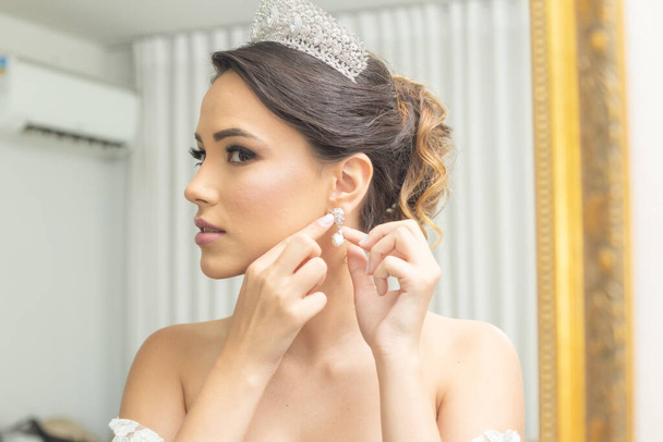 Young woman in a wedding dress, in front of the mirror, putting on her earrings. - Foto, Imagen