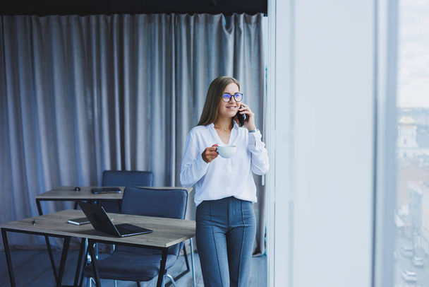 Фото успешной молодой женщины в рубашке, улыбающейся и работающей на ноутбуке, разговаривающей по телефону в современном офисе с большими окнами. Удаленная работа с кофе - Фото, изображение