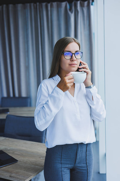 Photo of a successful young woman in a shirt smiling and working on a laptop while talking on the phone in a modern office with large windows. Remote work with coffee - Foto, afbeelding