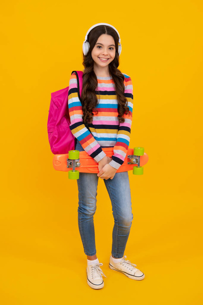 Teen school girl with skateboard backpack and headphone on studio isolated background. Schoolgirl trend, urban teenager style. Happy teenager, positive and smiling emotions of teen girl - Photo, Image
