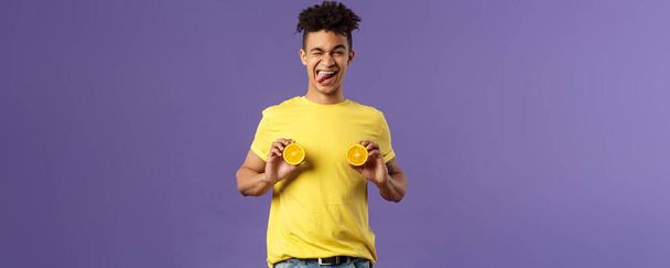 Holidays, vitamins and vacation concept. Portrait of sassy and cheeky funny young playful guy, fool around with fruit, holding pieces of oranges like women breast, show tongue smiling. - Photo, Image