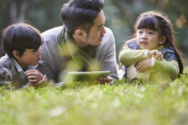 asiatischer Vater liegt vorne auf Gras und erzählt zwei Kindern fröhlich und lächelnd die Geschichte - Foto, Bild