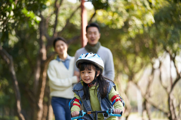 little asian girl riding bike in city park with parents in background - Foto, Imagen