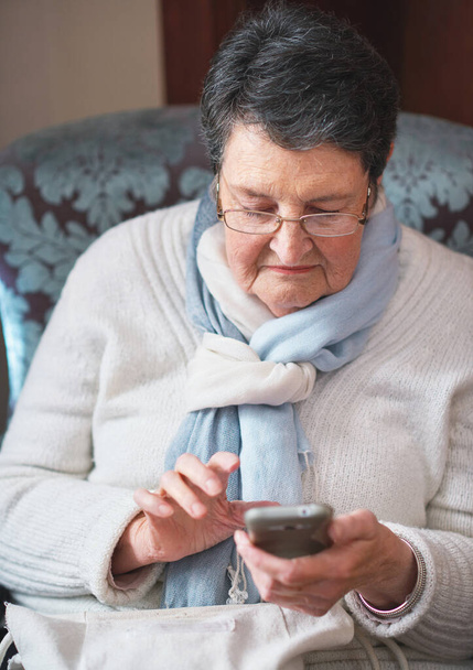 Happy elderly woman using smartphone texting browsing on mobile phone sending messages sitting on couch at home. - 写真・画像