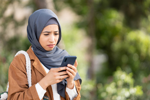 Μουσουλμάνα γυναίκα με μαντίλα χρησιμοποιεί το κινητό τηλέφωνο με μια θλιβερή έκφραση αηδίας σε ένα πάρκο - Φωτογραφία, εικόνα