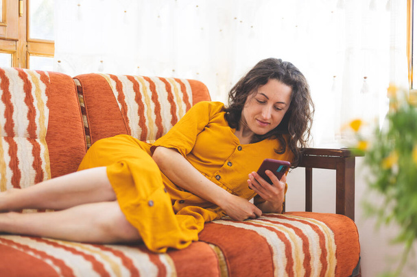 Дівчина в жовтій сукні лежить на дивані і тримає в руках смартфон, жінка відпочиває вдома і читає повідомлення
 - Фото, зображення