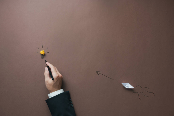 Conceptual image of business vision, innovation and startup - hand of a businessman drawing a light bulb with an origami made paper boat sailing towards it. Over brown background. - Foto, Imagen