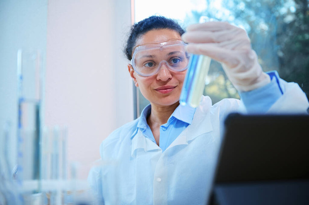 Close-up of beautiful African American woman, scientist, pharmacologist, clinical researcher wearing safety goggles, examining test tubes with chemical substances, in a science biological laboratory - Photo, image