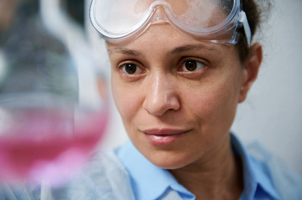 Headshot of confident experienced multi-ethnic woman, scientist chemist, pharmacologist inspecting the chemical reaction taking place in a glass flask with pink chemical fluid substance in laboratory - Photo, image
