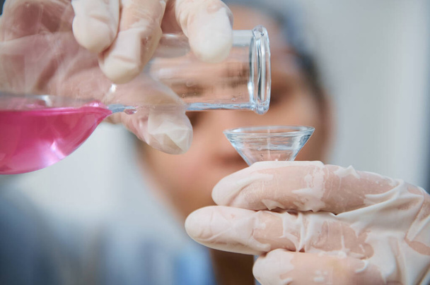 Подробности: Руки размытого сотрудника лаборатории, сливающего розовое жидкое химическое вещество из стеклянной пробирки через воронку в пробирку при проведении анализов в медицинской лаборатории - Фото, изображение