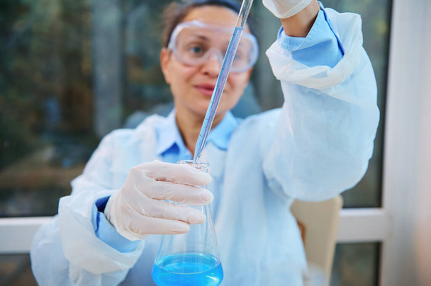 Focus on laboratory glass pipette in the hands of blurred woman scientist, medical biologist, researcher in a white lab coat, safety goggles and gloves pipetting blue liquid into a flat-bottomed flask - Zdjęcie, obraz