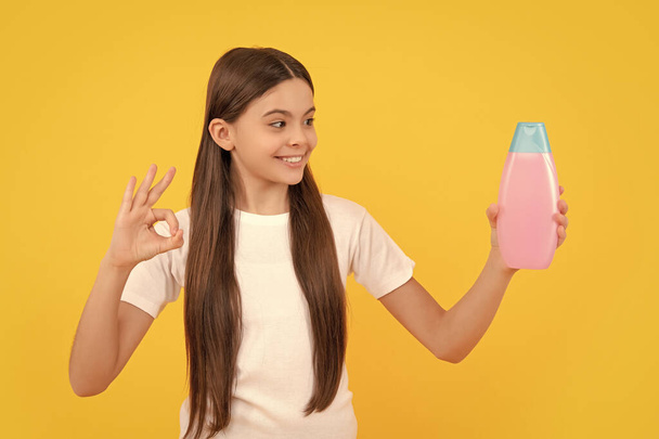 glücklich Kind halten Shampoo-Flasche zeigen ok Geste auf gelbem Hintergrund, Hautpflege. - Foto, Bild