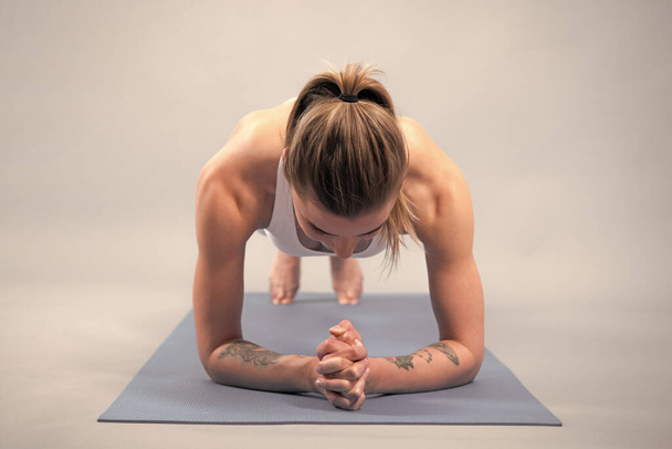 Een jonge atletische vrouw die planken doet. Full length fitnessvrouw in witte sportkleding zittend op gymmat en yoga oefenend, geïsoleerd op grijze achtergrond - Foto, afbeelding