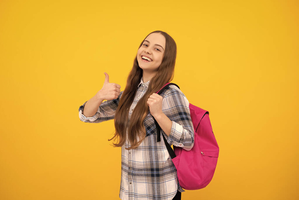 happy teen girl in checkered shirt carry backpack showing thumb up on yellow background, school. - Zdjęcie, obraz