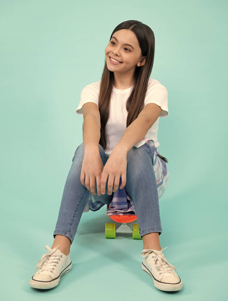Happy vogue teenager. Fashionable confident teenage girl wearing trendy t-shirt and jeans sit on skateboard, posing on blue background. Fashion teenager, casual look - Photo, image