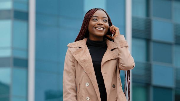 Young positive pretty african american woman standing against background of office building talking on phone answering mobile friendly call remotely chatting with friend using smartphone laughing - Photo, Image