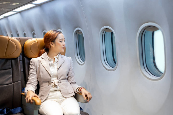 Ασιάτισσα γυναίκα ταξιδεύει κάθεται άνετα στο αεροσκάφος, ενώ κοιτάζοντας έξω από το παράθυρο για επαγγελματικά ταξίδια στην οικονομική θέση - Φωτογραφία, εικόνα