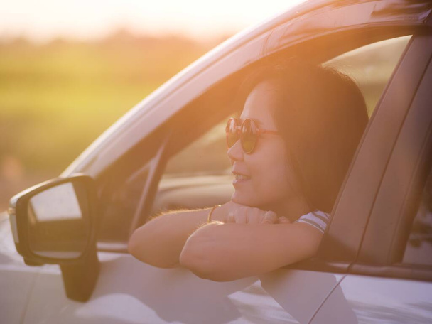 Όμορφη χαμογελαστή Ασιάτισσα γυναίκα με γυαλιά ηλίου κοιτάζοντας έξω από το παράθυρο λευκό αυτοκίνητο στο ηλιοβασίλεμα στο γήπεδο στη φύση.Ταξίδια και διακοπές έννοια - Φωτογραφία, εικόνα