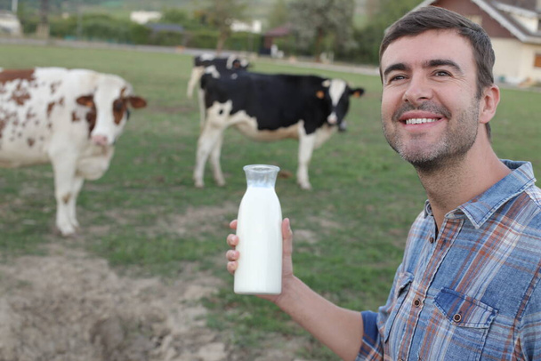 Porträt eines hübschen jungen Mannes, der mit einer Flasche Milch auf einer grünen Wiese steht, mit grasenden Kühen im Hintergrund - Foto, Bild