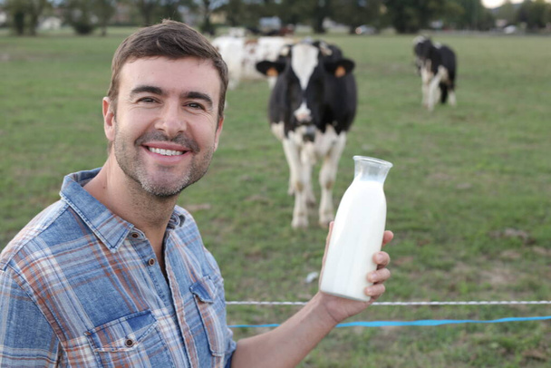 portret van knappe jongeman met flesje melk op groene meadfow met grazende koeien op achtergrond - Foto, afbeelding