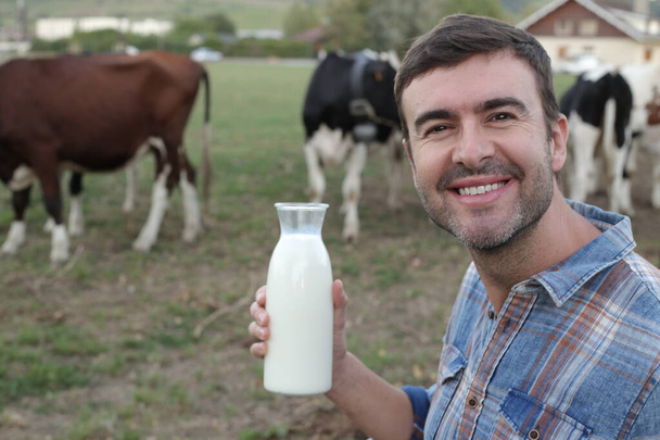 portrait d'un beau jeune homme debout avec une bouteille de lait sur une prairie verte avec des vaches brouteuses sur fond - Photo, image