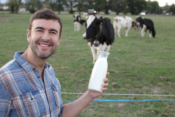 portret van knappe jongeman met flesje melk op groene meadfow met grazende koeien op achtergrond - Foto, afbeelding