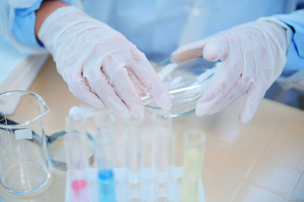 Selective focus on the hands in protective medical gloves of a lab assistant, laboratory technician, pharmacologist pouring white powdery substance from a test tube into a Petri dish during experiment - Foto, Imagen