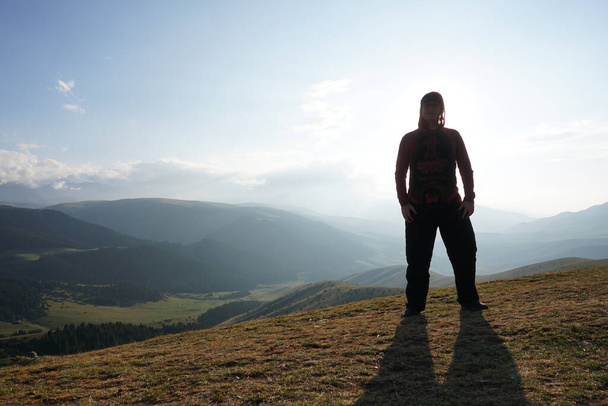 A guy on the edge of the hill admires the view. In the distance you can see high mountains and green hills covered with forest. Wide margins. Clouds are visible and sun is shining brightly - Foto, afbeelding