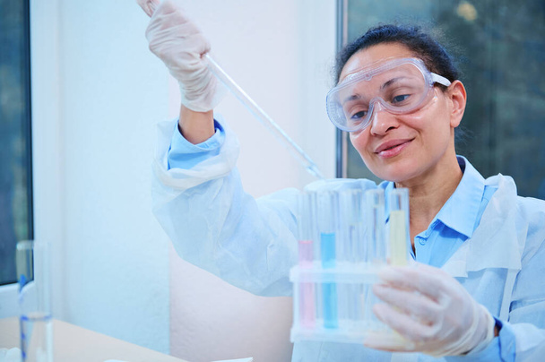 Inspired Latin American woman, lab assistant, scientist chemist using a glass dropper, dripping few chemical reagent into test tubes, while conducting experiment in a clinical research laboratory - Photo, image